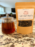 Tea bag packaging labeled 'Soft Start Black Tea' next to a glass teapot with tea on a marble surface.